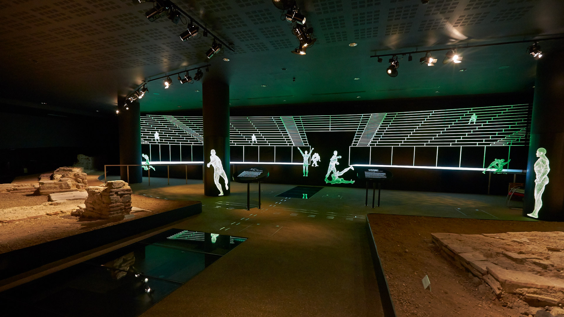 Family Fun Activities to Enjoy on the Weekend in the City of London - Roman Ampitheatre Family Fun Activities to Enjoy on the Weekend in the City of London - Roman ruins of an amphitheatre in a dark room with lit figures around the room doing traditionally roman sports actvities