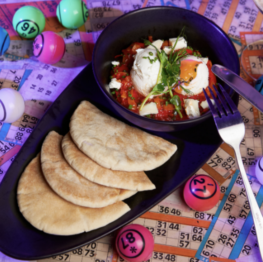 9 Best Brunch Spots in the City of London - Dabbers Bingo 9 Best Brunch Spots in the City of London - Dabbers Bingo - table covered in bingo cards and balls with a plate of pitta and a bowl of Shakshouka and poached eggs