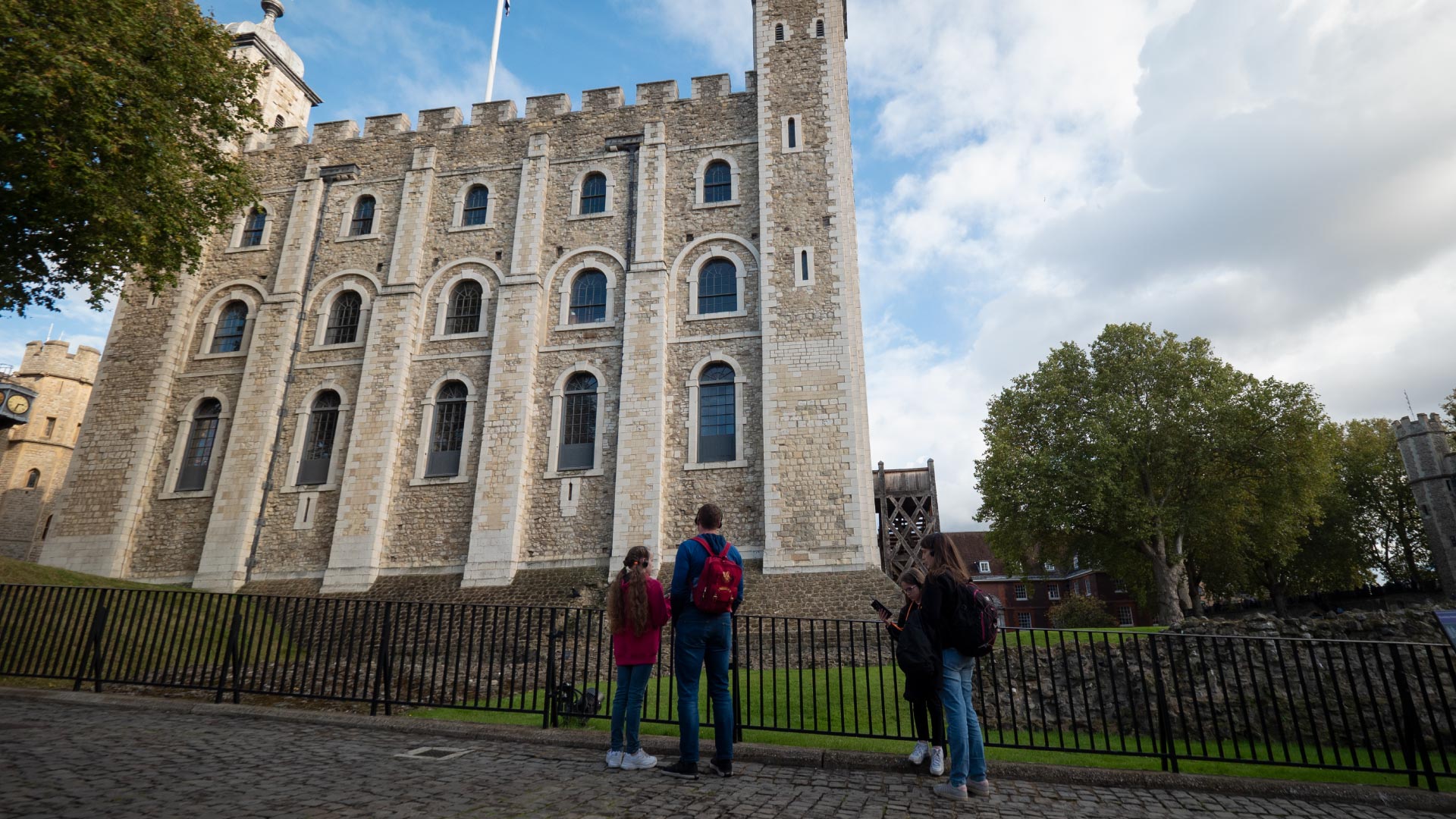 Family Fun Activities to Enjoy on the Weekend in the City of London - Tower of London Family Fun Activities to Enjoy on the Weekend in the City of London - A picture of the Tower of London on a summer day, behind the fence there is a family of four standing and listening to an audio guide, an older male, and thee younger girls.