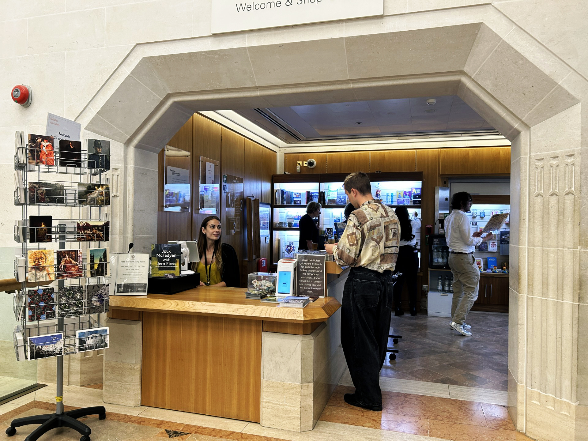 A visitor interacts with a staff member at the Guildhall Art Gallery gift shop counter. Nearby, others browse items on shelves, surrounded by historical memorabilia. A display rack stands by the entrance.