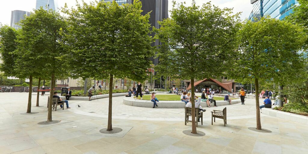 Aldgate Square in the City of London. Green trees, a lawn and people sitting and enjoying the sunshine.
