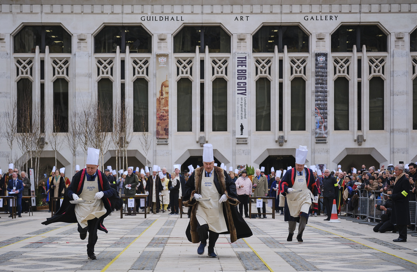 Three people where chef hats and robes running in Guildhall Yard holding a frying pan as part of the annul inter-livery pancake day race.
