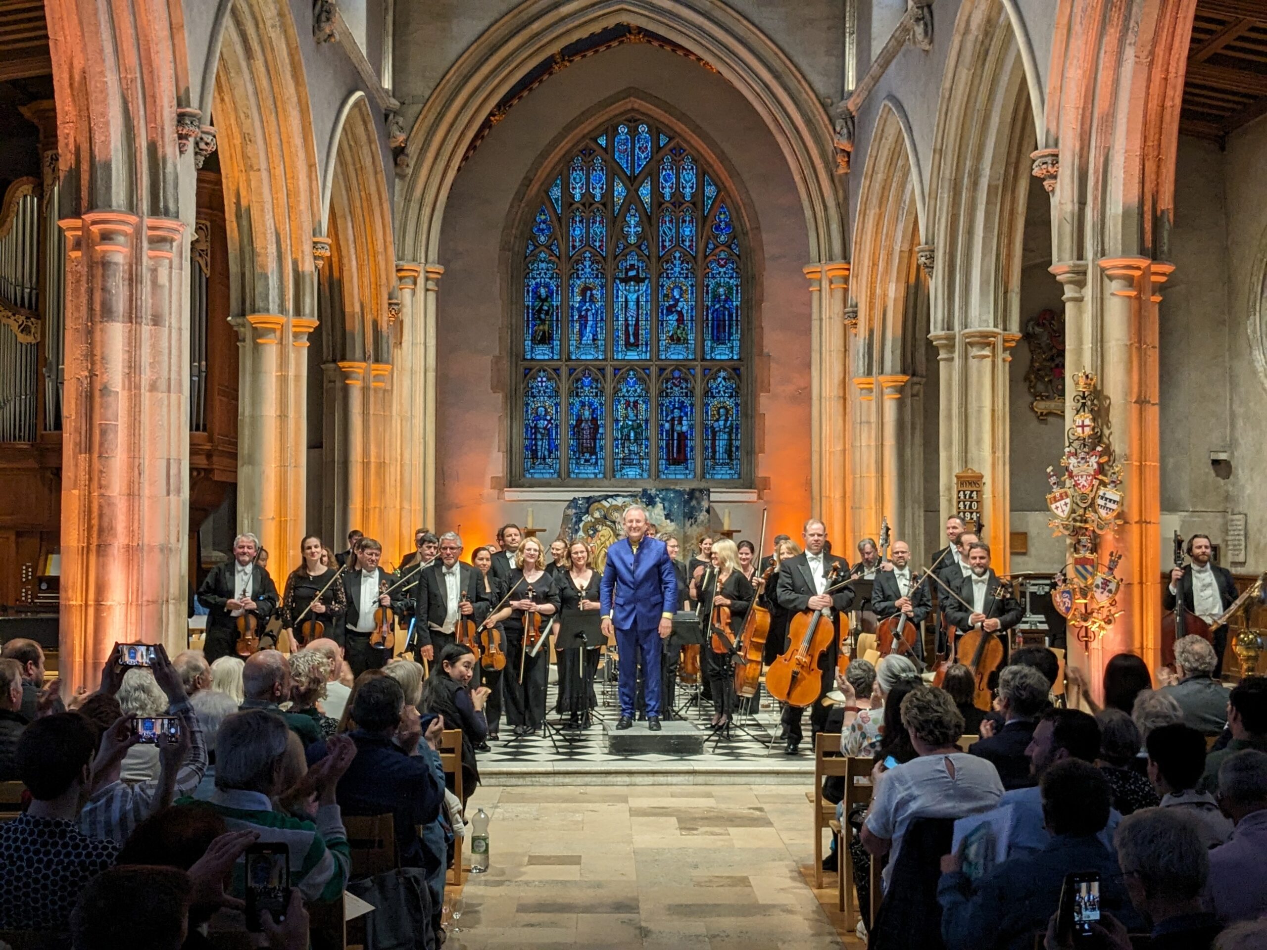 A man in a vibrant blue suit stands in a church facing a seated audience with a orchestra standing behind him.