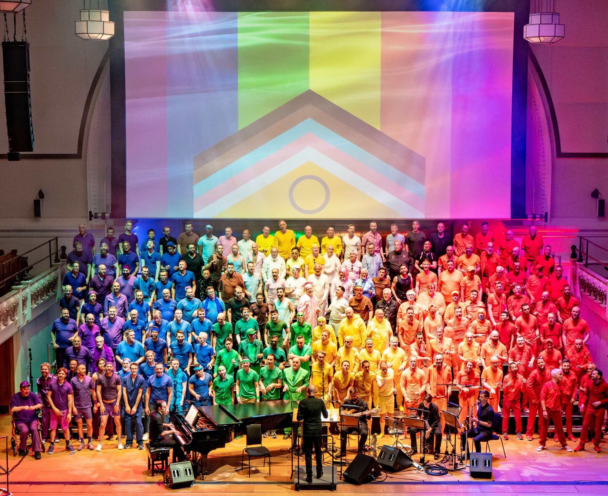 A large group of peope on stage with sections wearing different coloured shirts representing the pride flag, with a large pride flag behind them