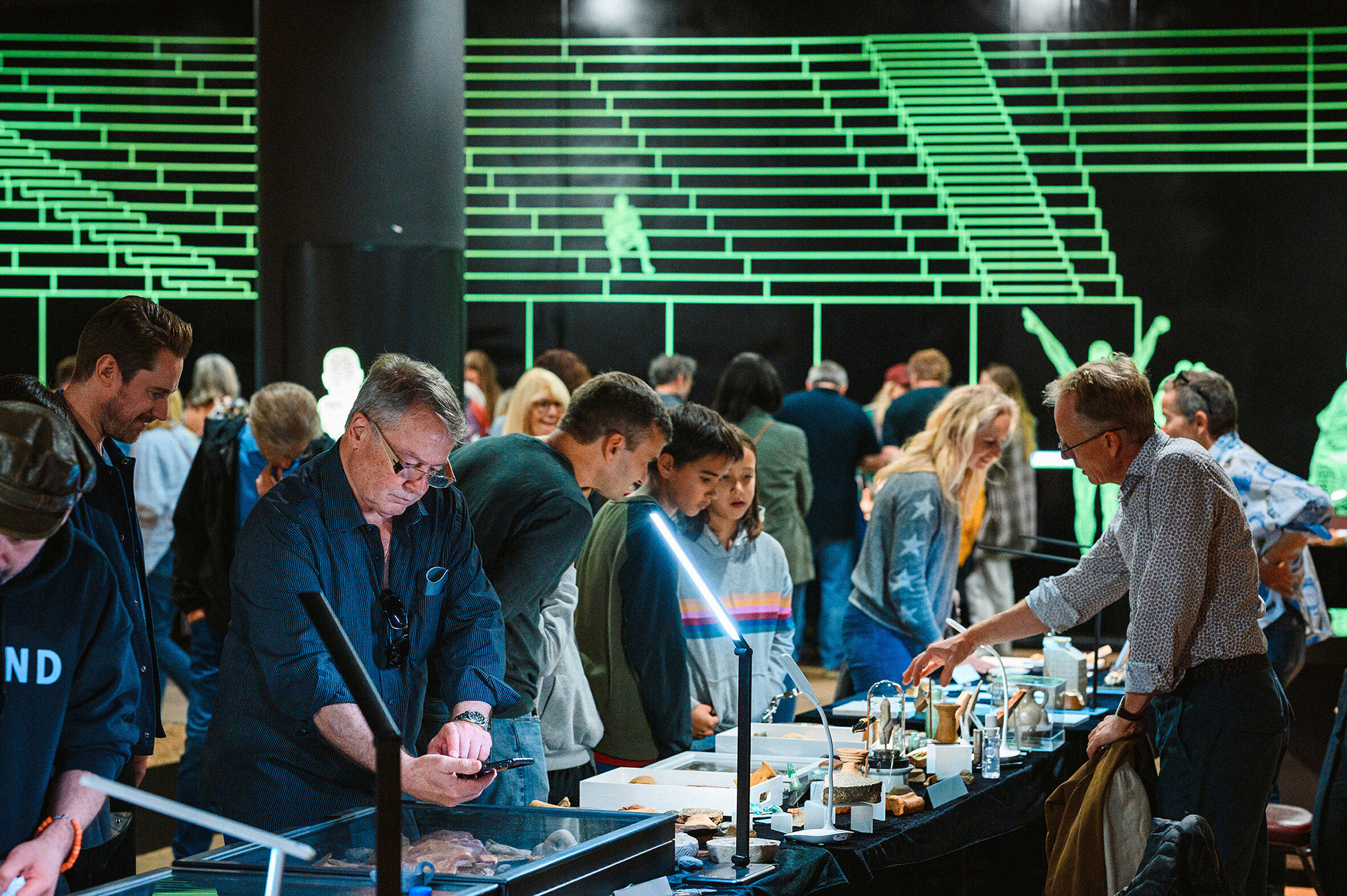 Visitors of various ages engage with interactive exhibits at London's Roman Amphitheatre, surrounded by bright neon green light installations.