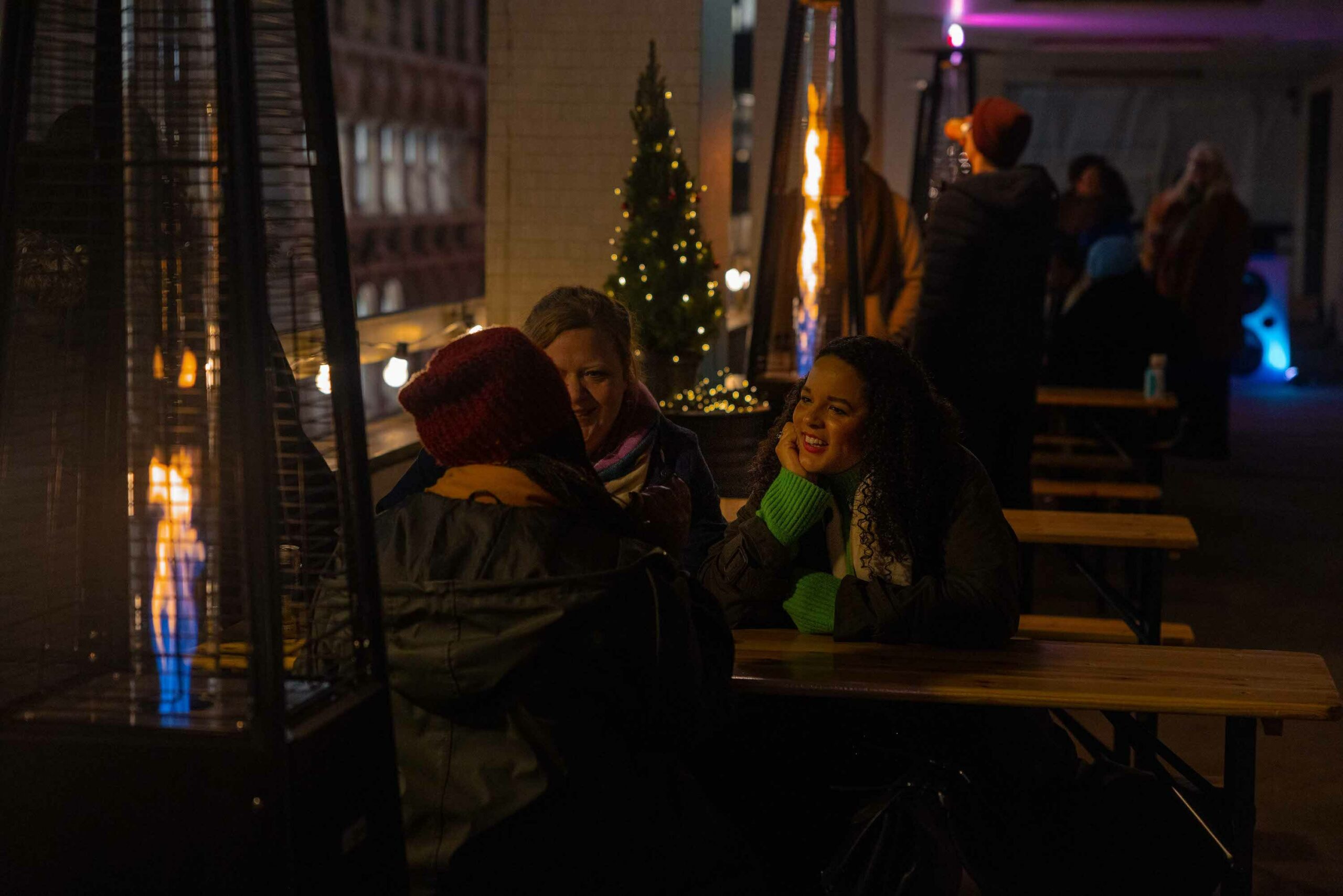 Group of people enjoying a conversation at an outdoor table with a heating lamp, surrounded by festive lights and decorations.