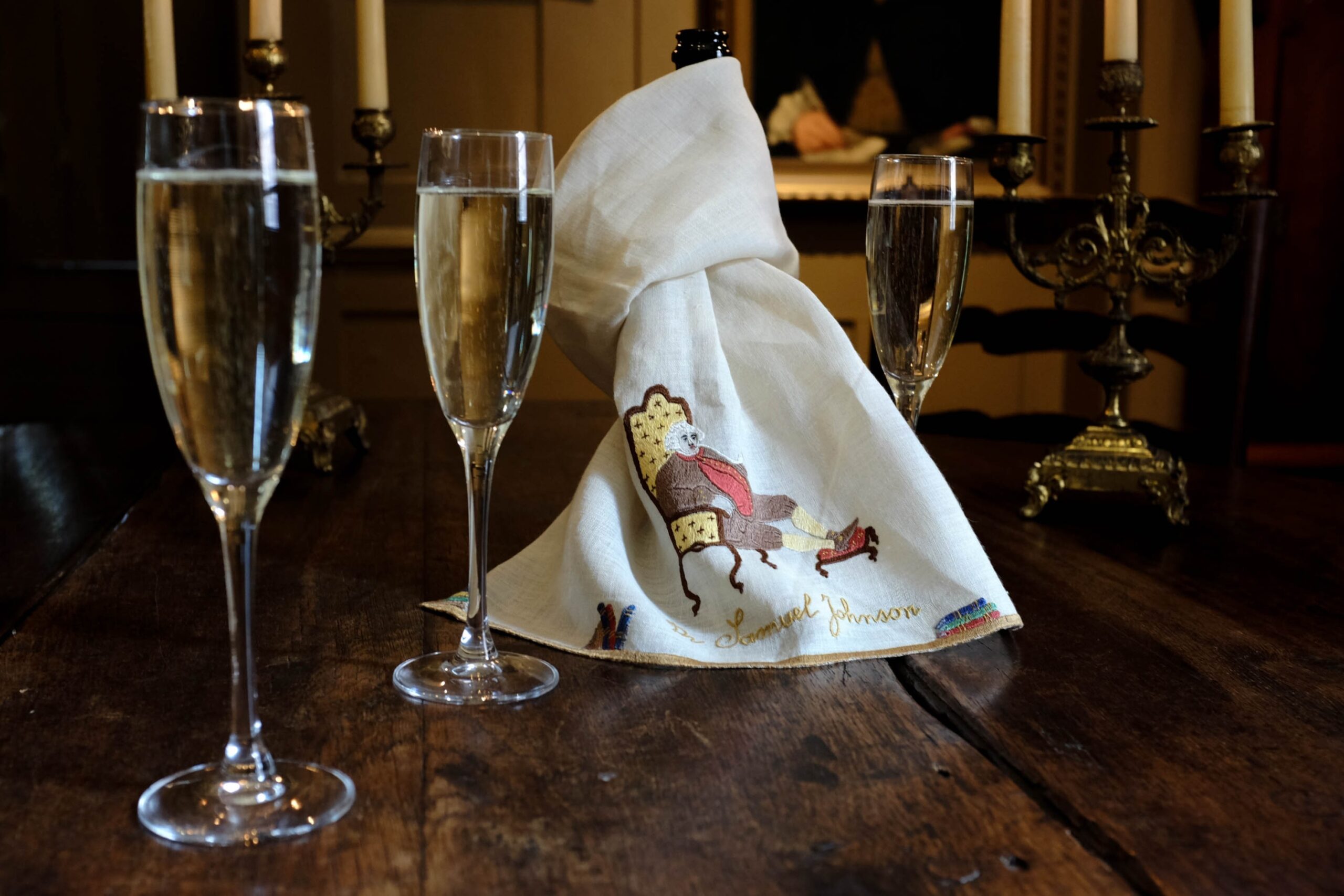 Two champagne glasses on a wooden table, with an intricately folded napkin featuring a embroidered image of Dr Johnson. In the background, a candlelit setting enhances the ambiance.