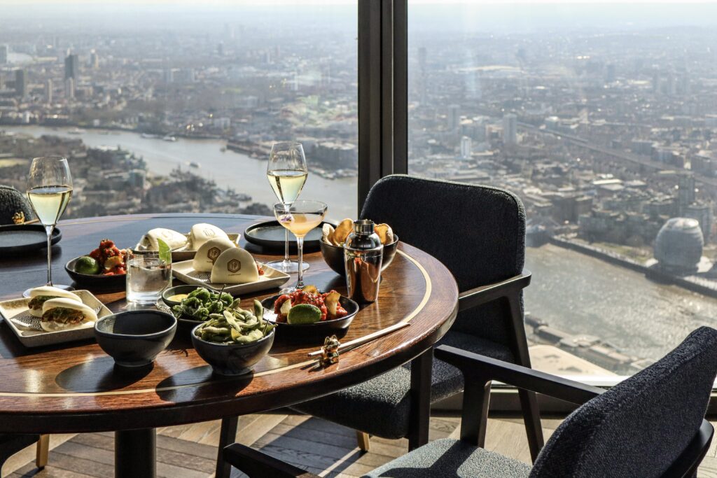 A table with a variety of dishes and glasses of wine, set near a large window overlooking a panoramic view of the Thames River and the London skyline.