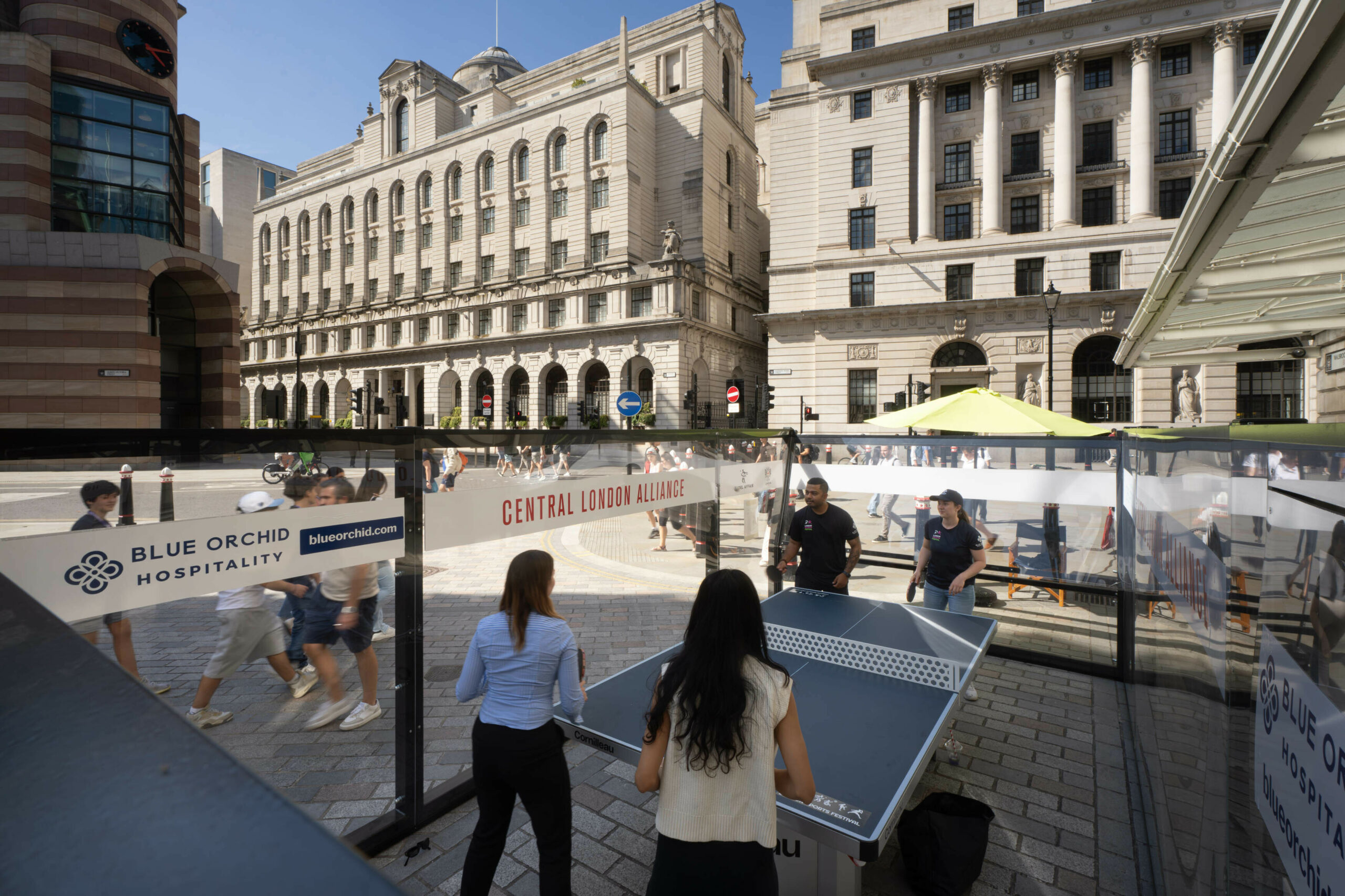 View of people engaging in a padel table tennis game outside Mansion House on a sunny day, with spectators watching from behind a transparent barrier displaying logos for Blue Orchid Hospitality and Central London Alliance.