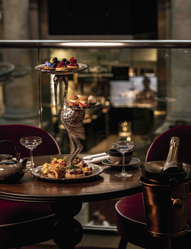 An elegant afternoon tea setup featuring a three-tiered serving stand shaped like a stork, adorned with desserts and pastries, on a table with champagne and a teapot, within a plush dining setting.