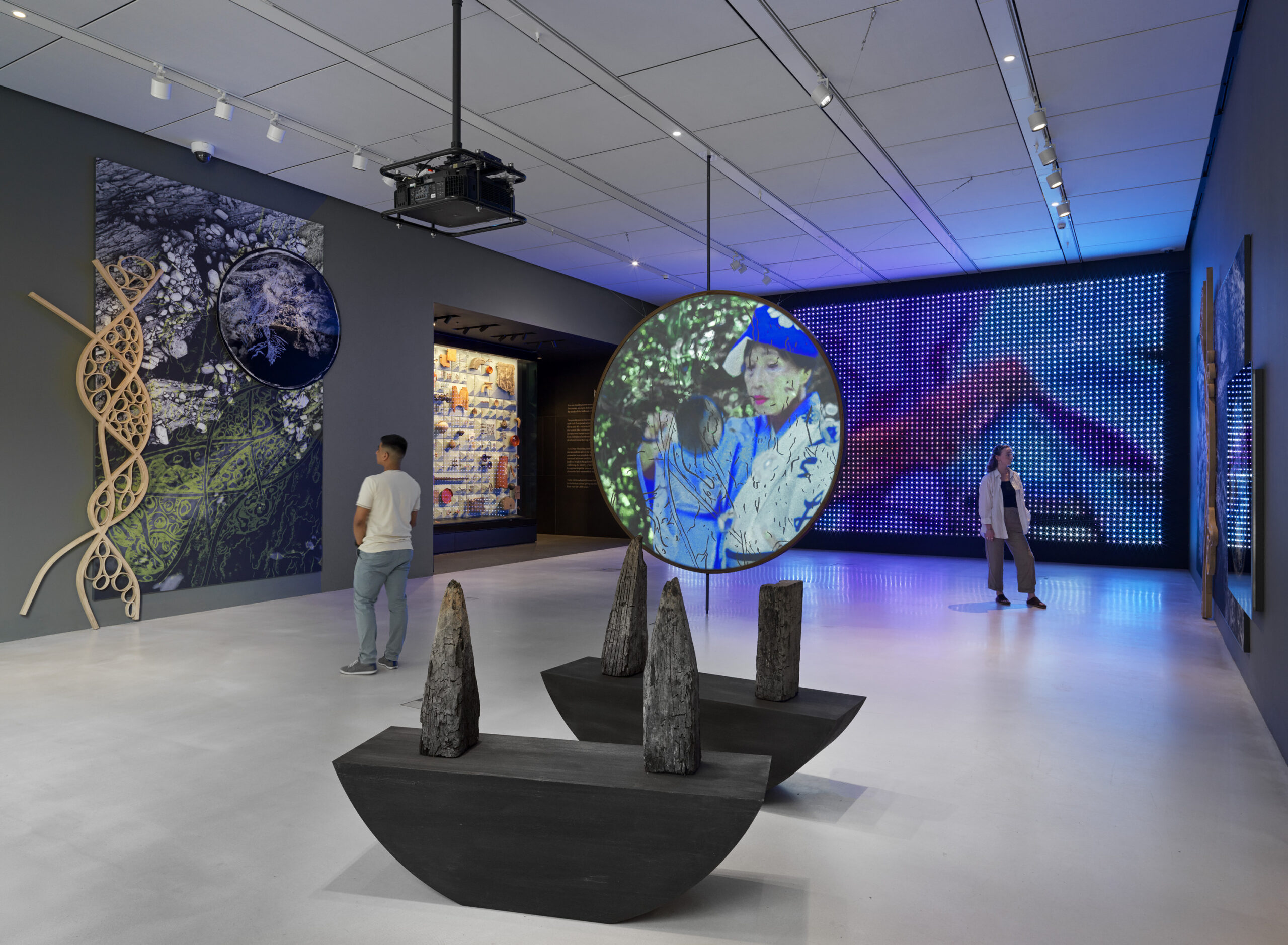 Visitors explore an exhibition featuring diverse multimedia displays, including a large circular screen projecting an image of a person, surrounded by abstract sculptures.