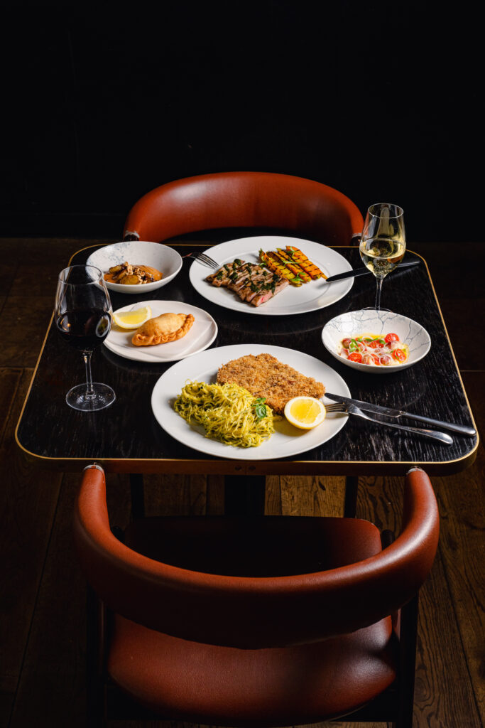 A well-arranged dinner table at a restaurant featuring multiple dishes including pasta, salad, grilled meat, and soup, complemented with a glass of white and red wine.