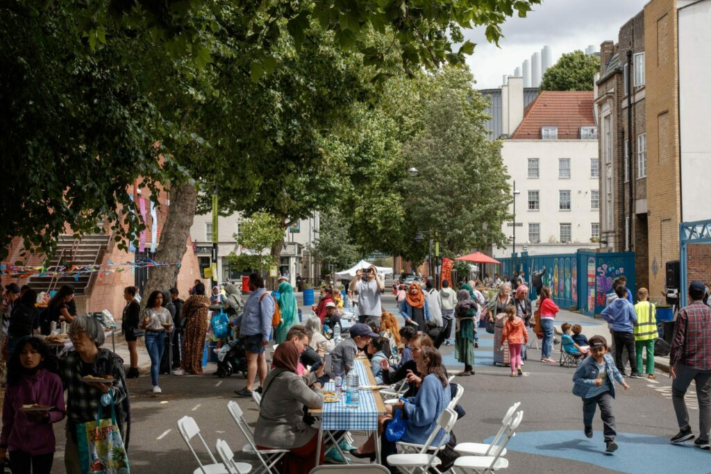 A vibrant street fair scene with people of various ages socialising and participating in activities like chess. Trees and urban buildings surround the area.
