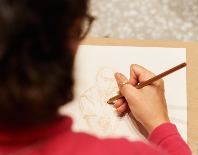An over-the-shoulder look at someone sketching a person. The drawer is wearing a pink turtleneck.