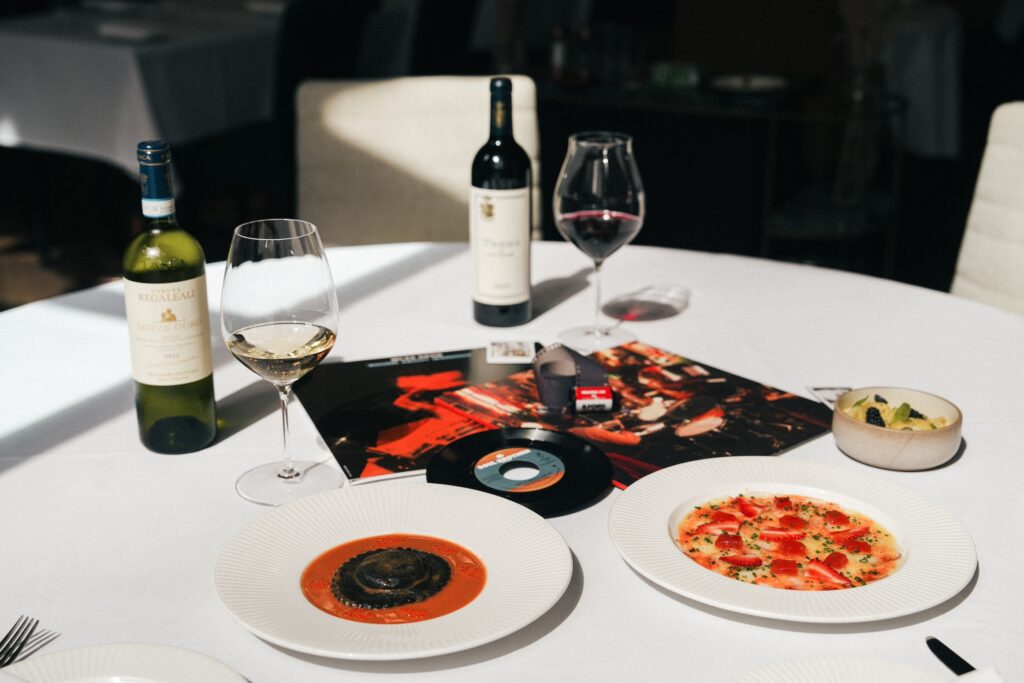 A table is set with two plates of gourmet food and two glasses of wine, one white and one red, beside a bottle of Montecillo wine. A vinyl record and an open booklet with a vibrant red design are also on the table, suggesting a themed dining experience.
