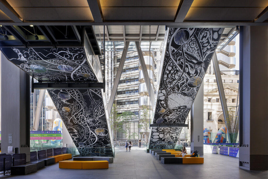 View under The Leadenhall Building, featuring the underside of two escalators with intricate black and white designs. People are sitting on colourful benches, enjoying the shaded outdoor area.