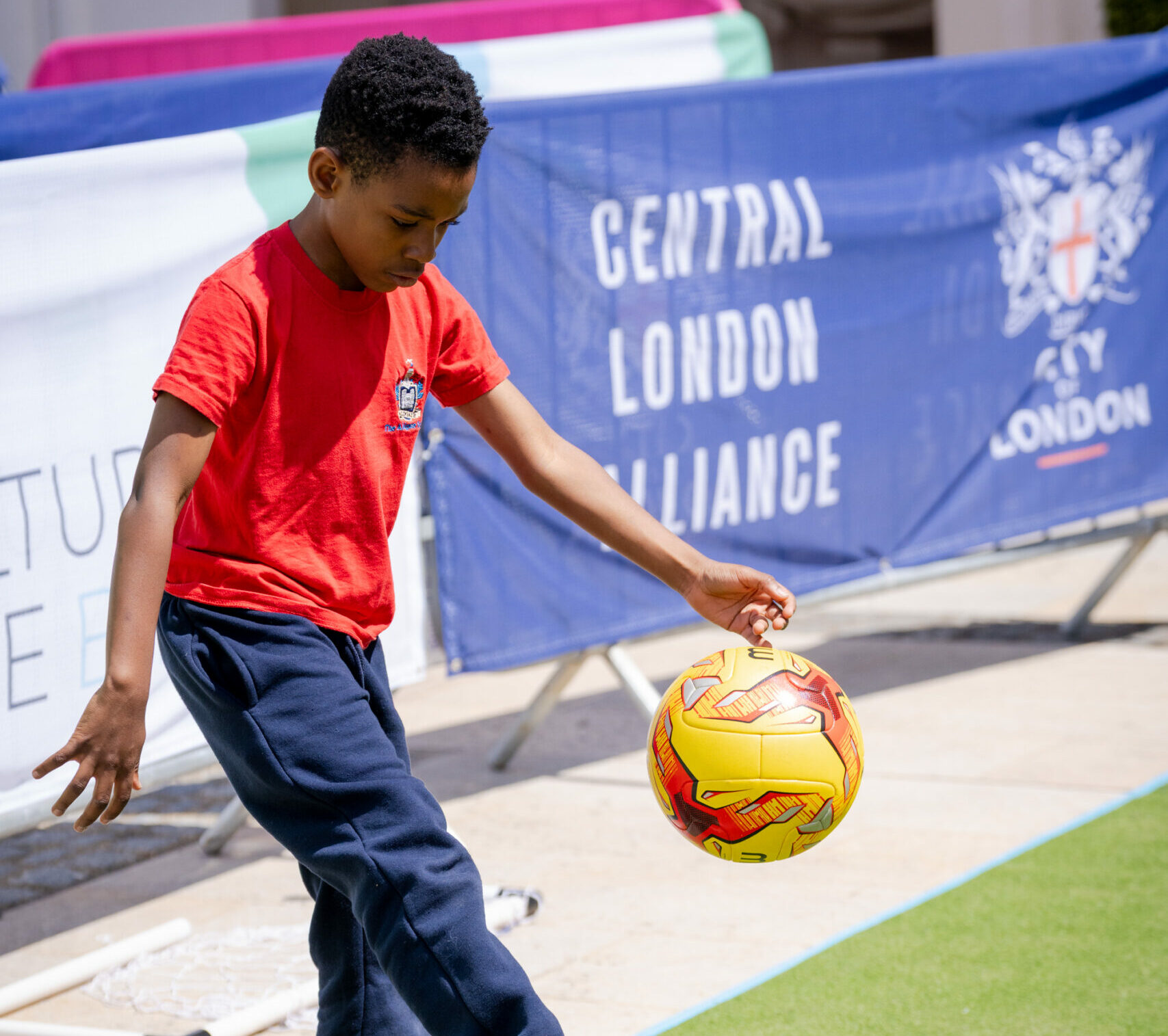 A child playing football on a small field with a banner reading "Central London Alliance" in the background.