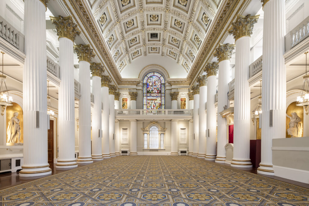 Interior view of the Egyptian Hall in Mansion House (City of London), featuring ornate ceilings, large pillars, statues, and a decorative floor with a stained-glass window at the far end.