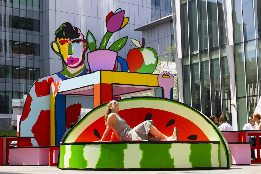 Person relaxing on a watermelon-shaped bench in front of a colourful sculpture featuring a stylized face and fruit, located in a vibrant urban square.