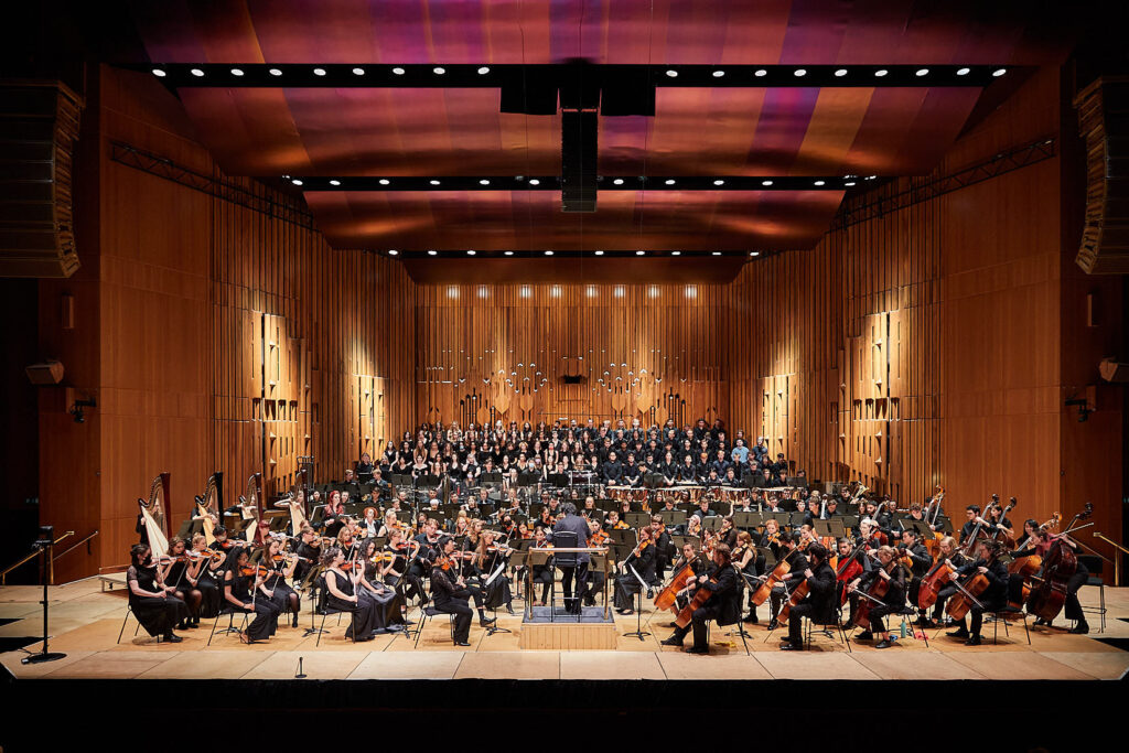 Wide view of Guildhall Symphony Orchestra & Chorus performing on stage in a concert hall with musicians seated in rows and a choir standing in the background.