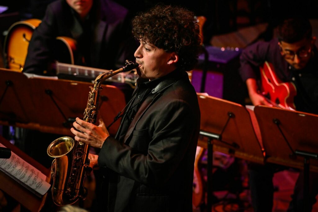 A musician playing a saxophone on stage at a live concert, accompanied by a guitarist and other band members in the background. The setting includes music stands and stage lighting.