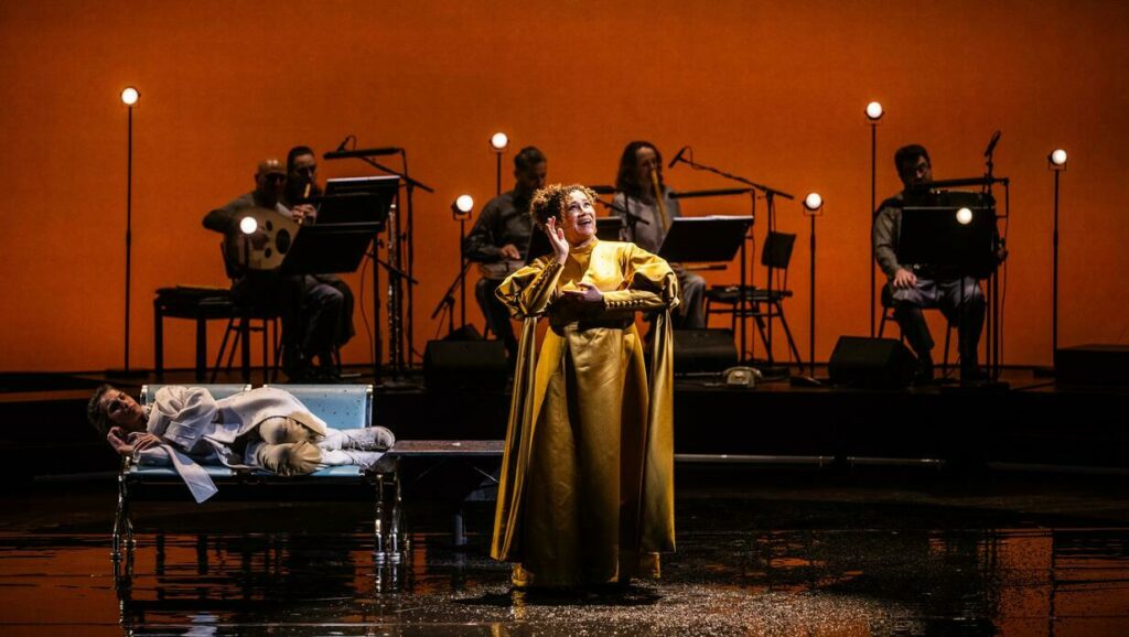 Performance of the opera 'Oum - A Son's Quest for his Mother' with two vocalists on stage, one lying on a horizontal platform and the other standing in a vibrant yellow robe, surrounded musicians with sheet music in the background.