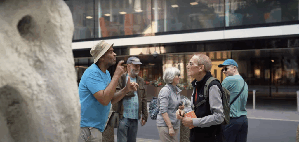 Two individuals engaging in a BSL conversation on a city street as part of a tur, with a bystander observing and a statue partially visible in the foreground.