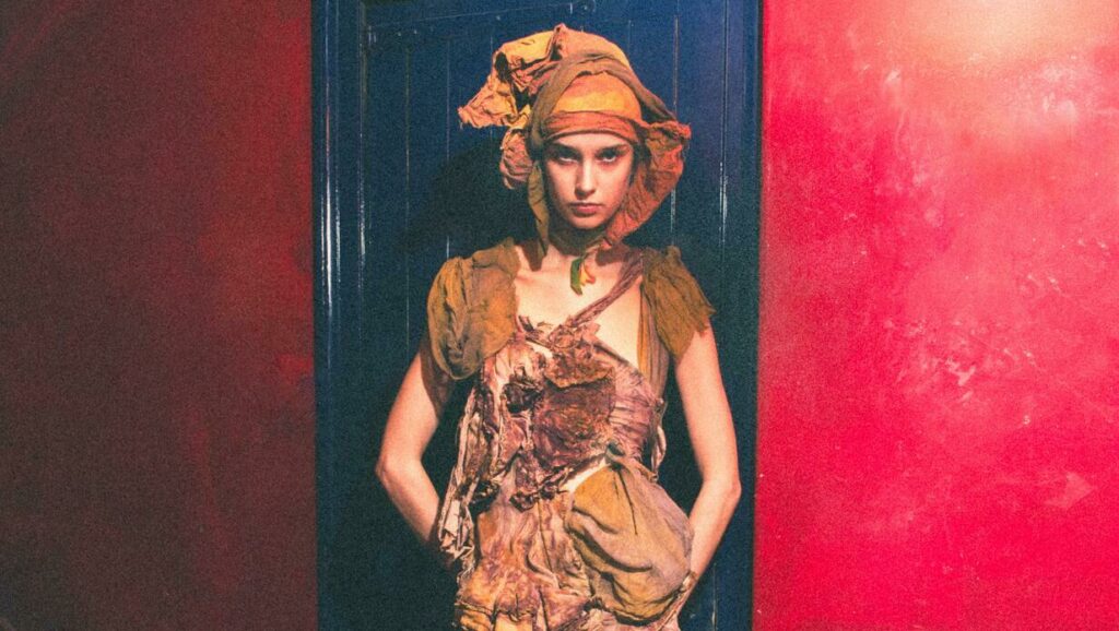 A person dressed in an elaborate, textured costume stands against a split background of dark blue and vibrant red.