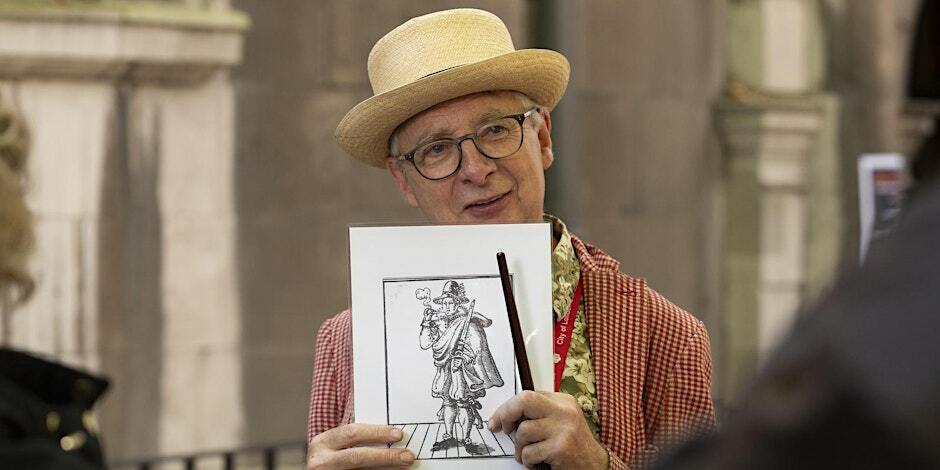 Person holding a framed sketch and a paintbrush, wearing a straw hat and spectacles, smiling at an event.