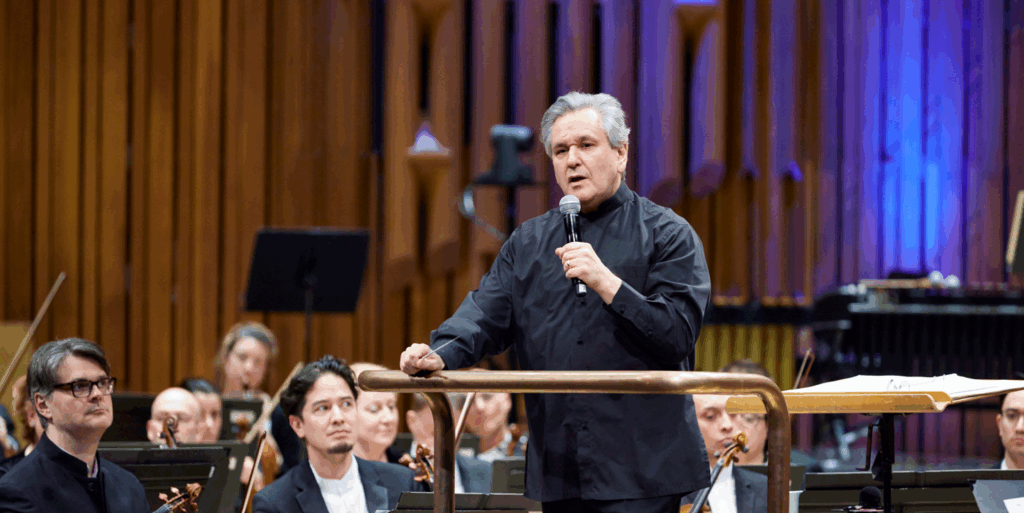 Conductor leading an orchestra during a performance, with violinists and other musicians in the background.