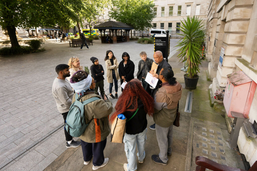 A diverse group of people engaged in a guided tour outdoors, listening attentively to a tour guide who is holding a pamphlet. The setting includes trees, benches, and a small kiosk in the background.