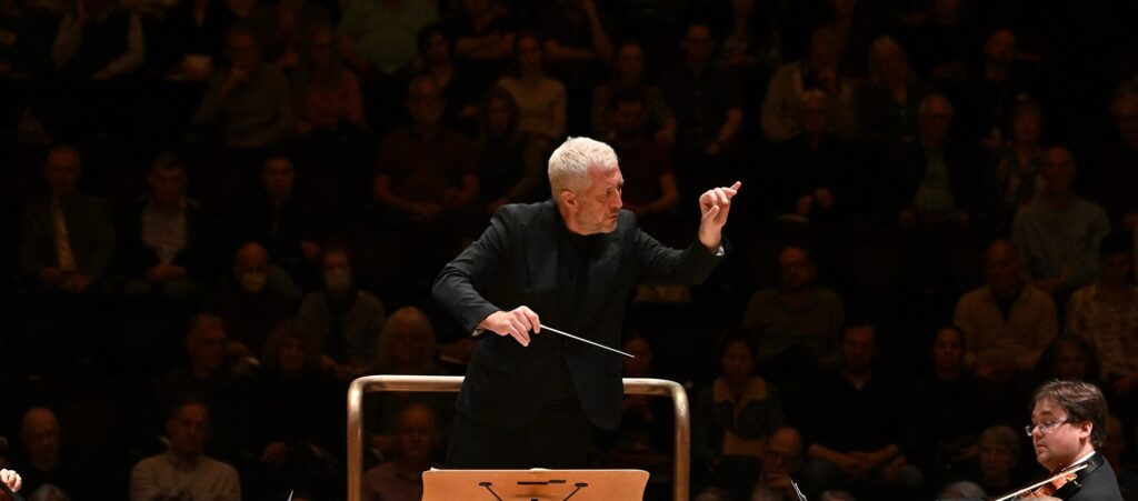 Conductor leading an orchestra during a live performance, gesturing expressively while facing the musicians.
