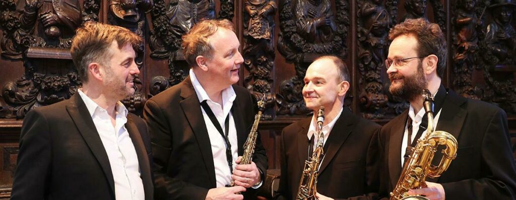 The four members of the Quartz Saxophone Quartet, each holding a saxophone, smiling and interacting joyfully on a stage with ornate wooden carvings in the background.
