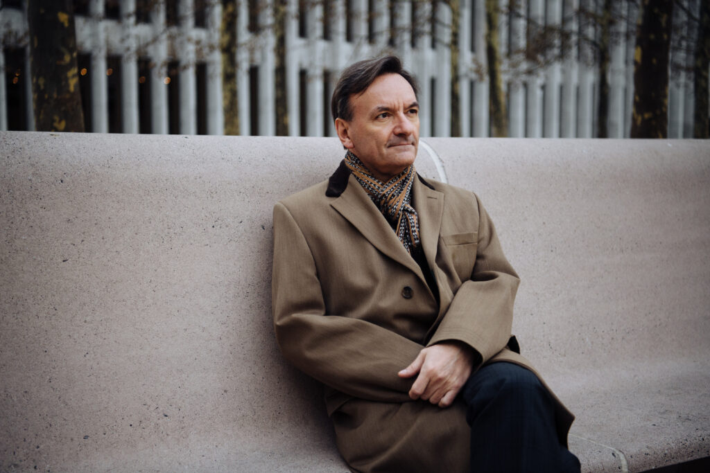 Stephen Hough sitting on a bench outdoors, wearing a coat and scarf, with a thoughtful expression. The backdrop features leafless trees and modern architecture.