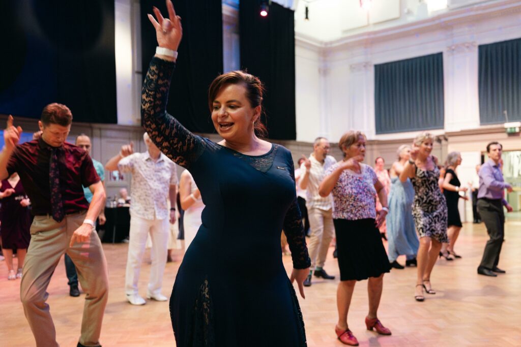 A person joyfully dancing in the forefront at a lively ballroom dance event.