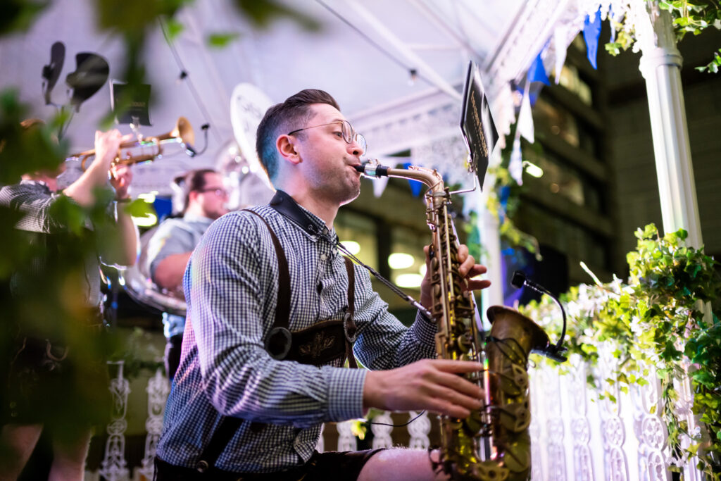A musician playing a saxophone, part of a live band performance in a vibrant setting with other band members in the background.