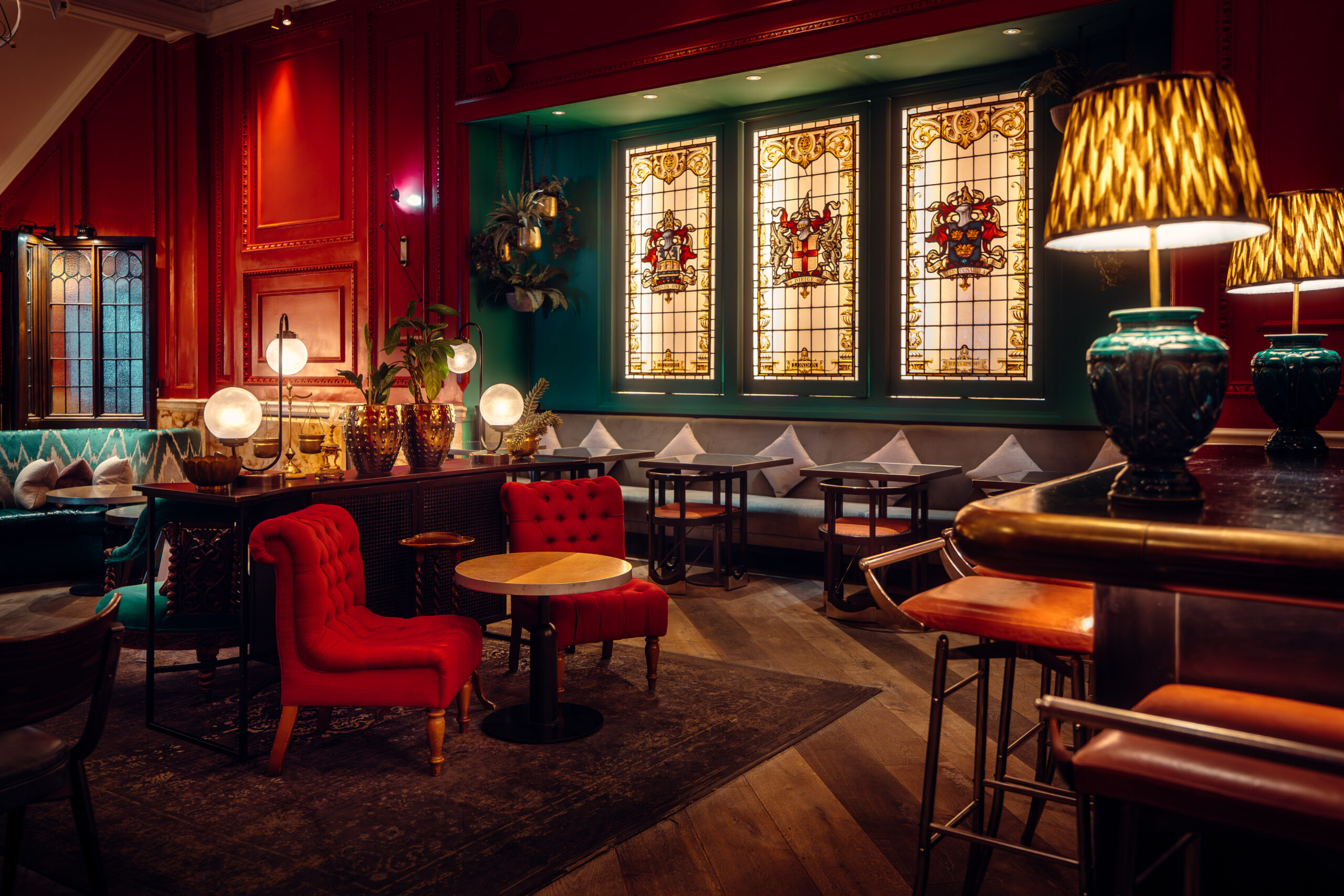 Interior view of a richly decorated restaurant with red walls, stained-glass windows, and various seating arrangements, including red upholstered chairs with small round tables and bar stools. Soft, warm lighting from lamps and fixtures.