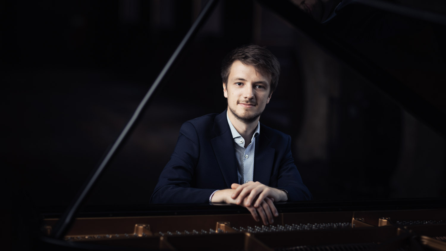 A man is facing the camera, resting his arms on a grand piano.