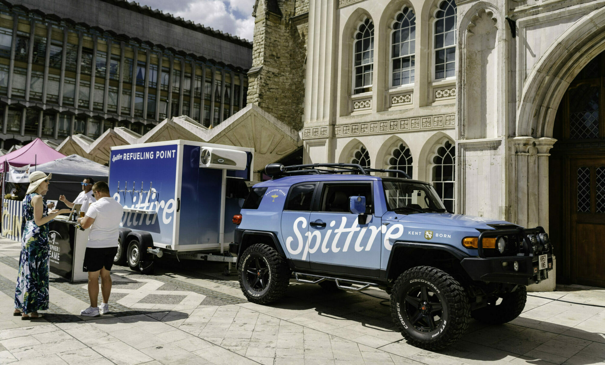 A branded blue car with a trailer is parked in a historic courtyard, with a group of people assembled behind the vehicle.