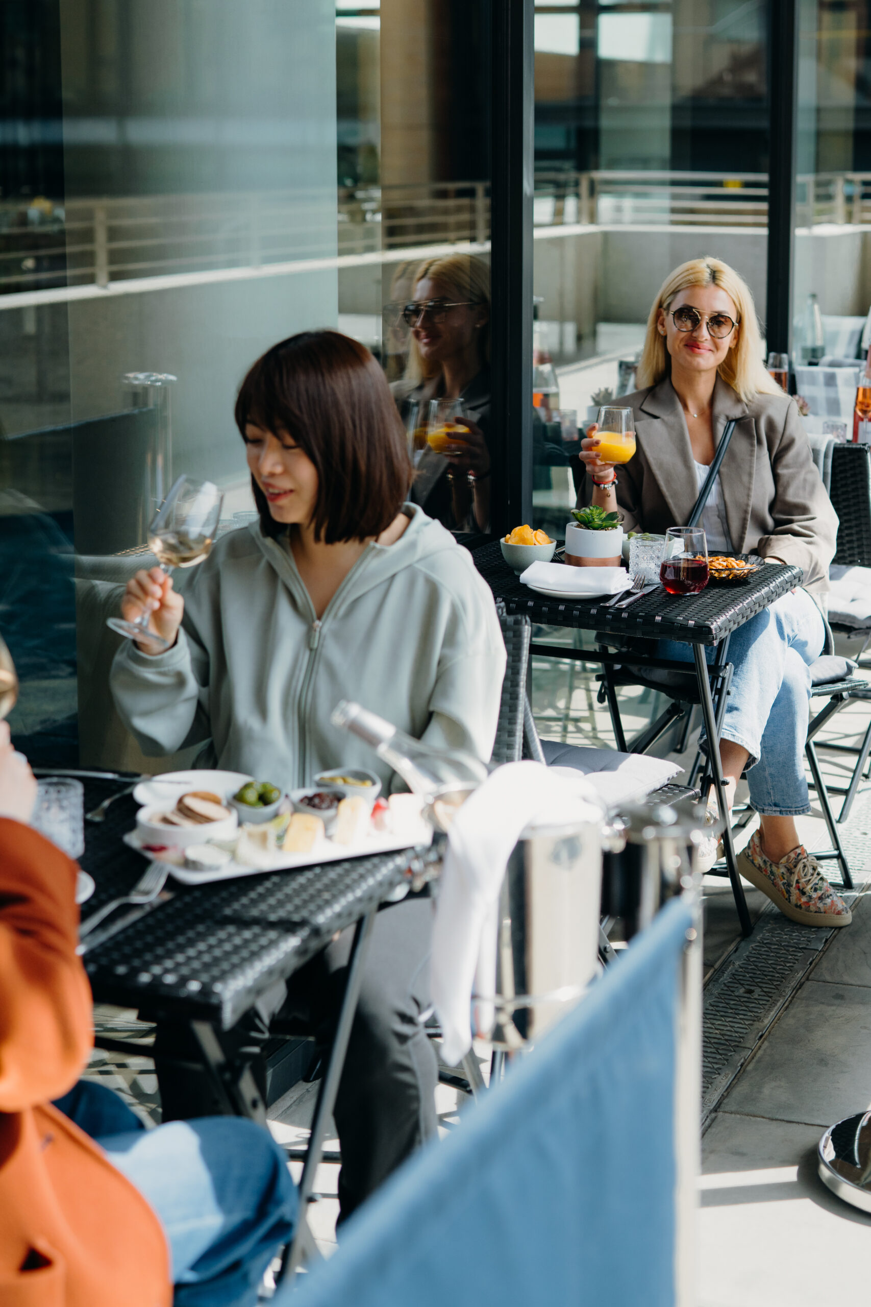 An image of two people sat at tables holding drinks outside, one in the background is wearing sunglasses