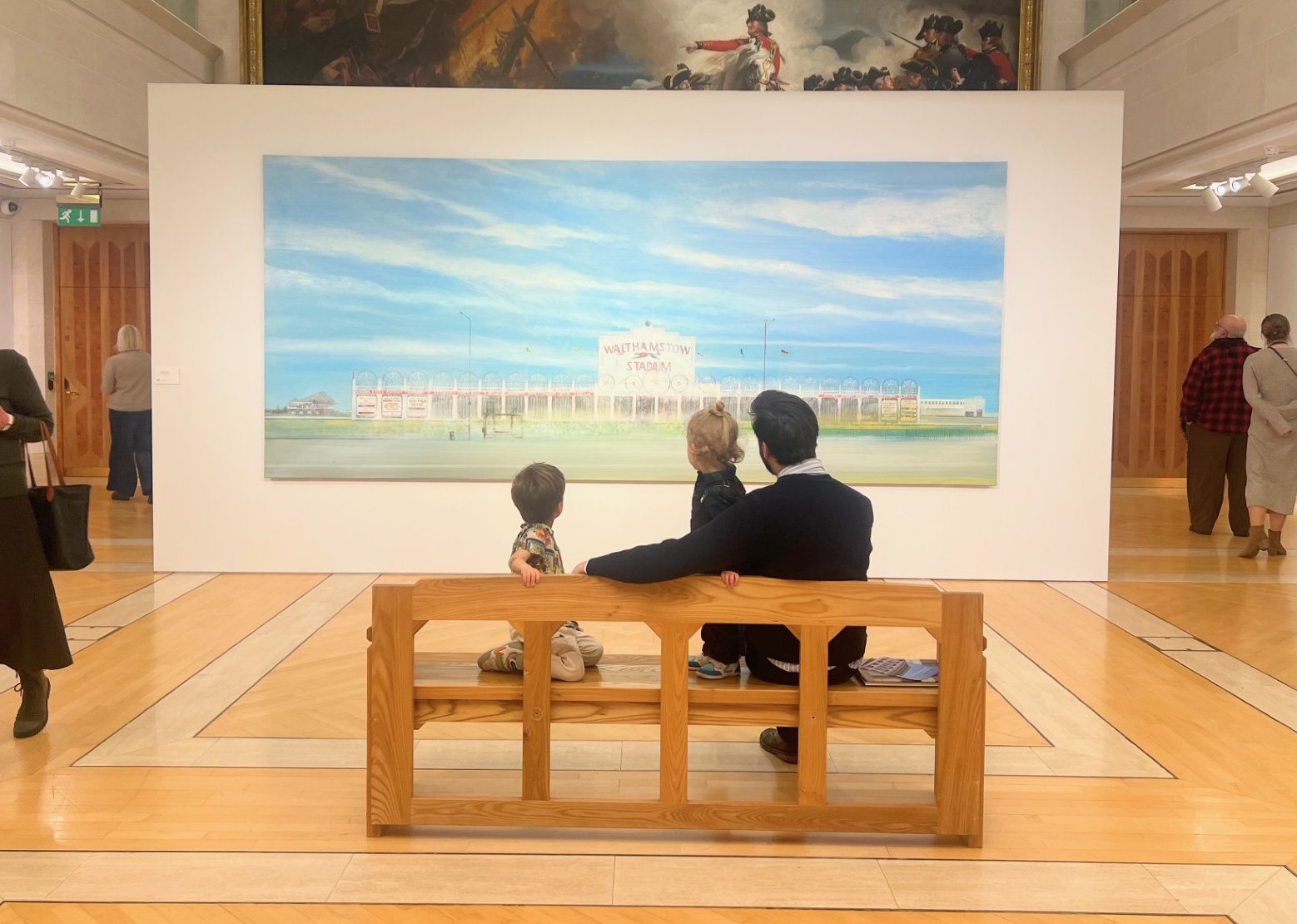 One adult and two children sitting on a bench looking at a large painting in a museum