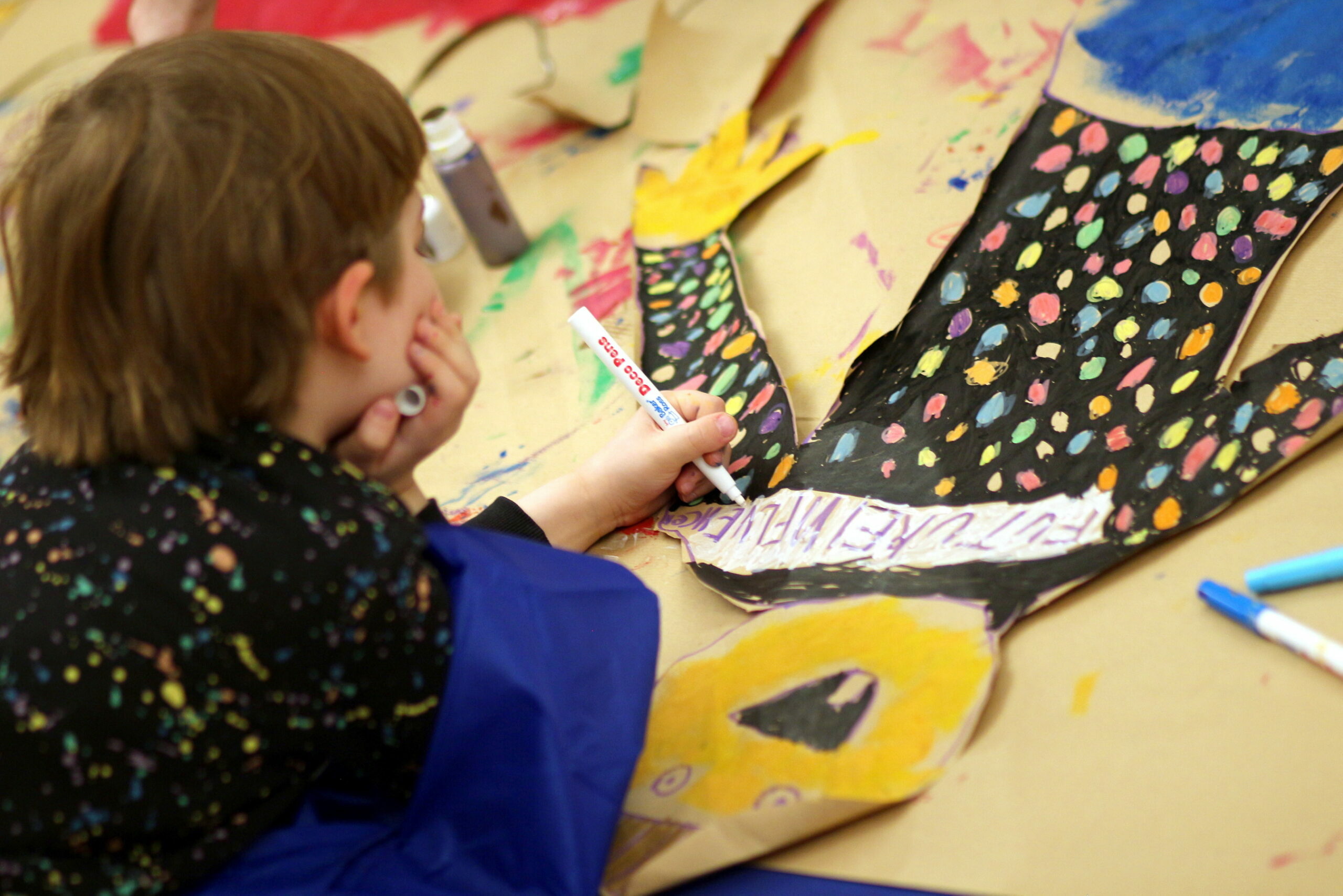 A person is drawing on a colorful, polka-dotted cutout of a glove. The background shows an art table with various markers and art supplies.