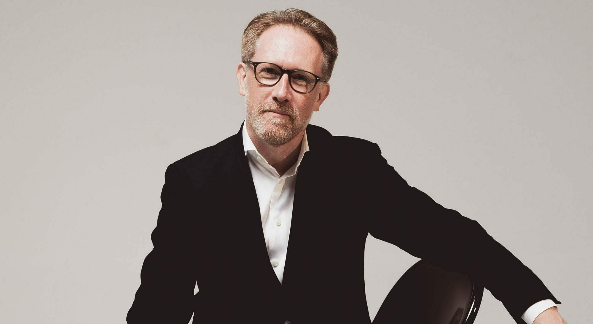 Portrait photo of music conductor, Simon Crawford-Phillips. Wearing a dark suit and sitting on a chair