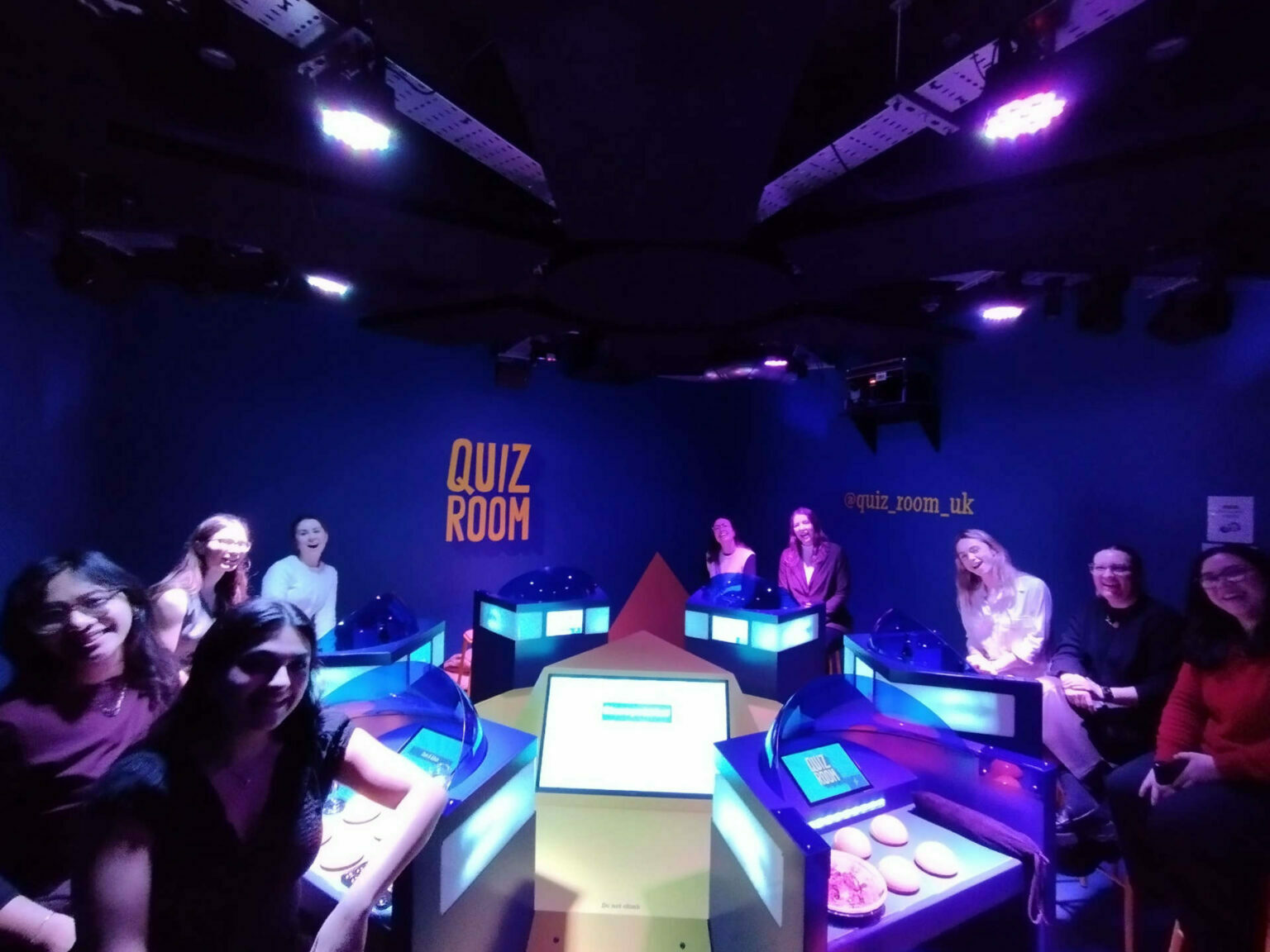 An image in a coloured lit up room with teams sat around a central console smiling for the cameras