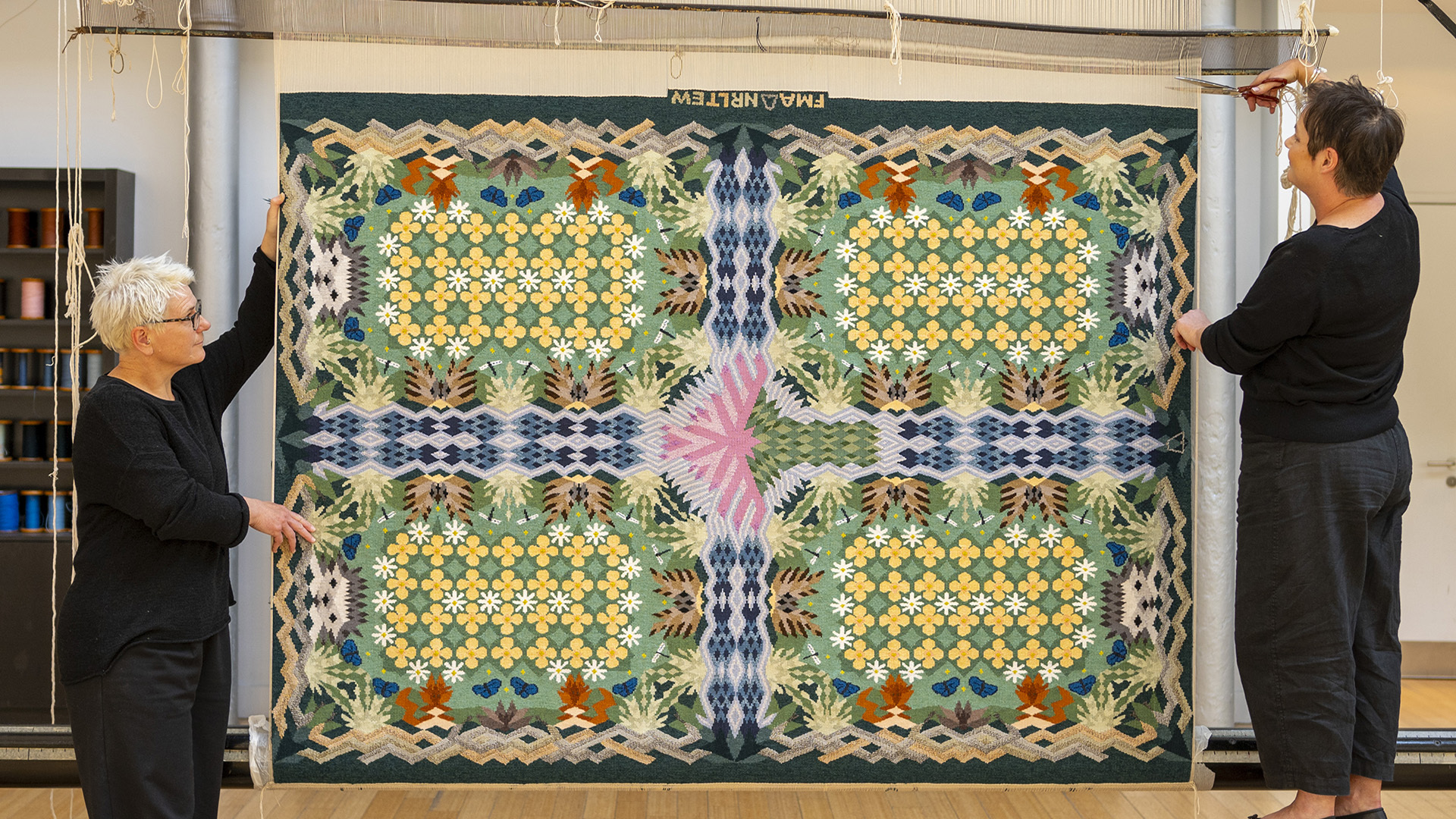 Two individuals hold up a large, colorful patterned rug with floral and geometric designs. 