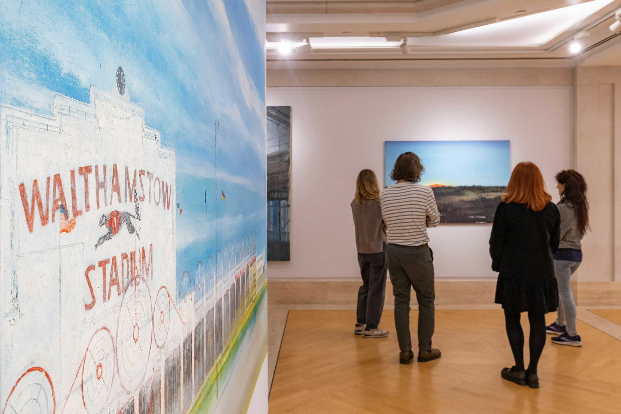 A group of people stand in an art gallery viewing paintings. One painting prominently features “Walthamstow Stadium” with a depiction of racing dogs.